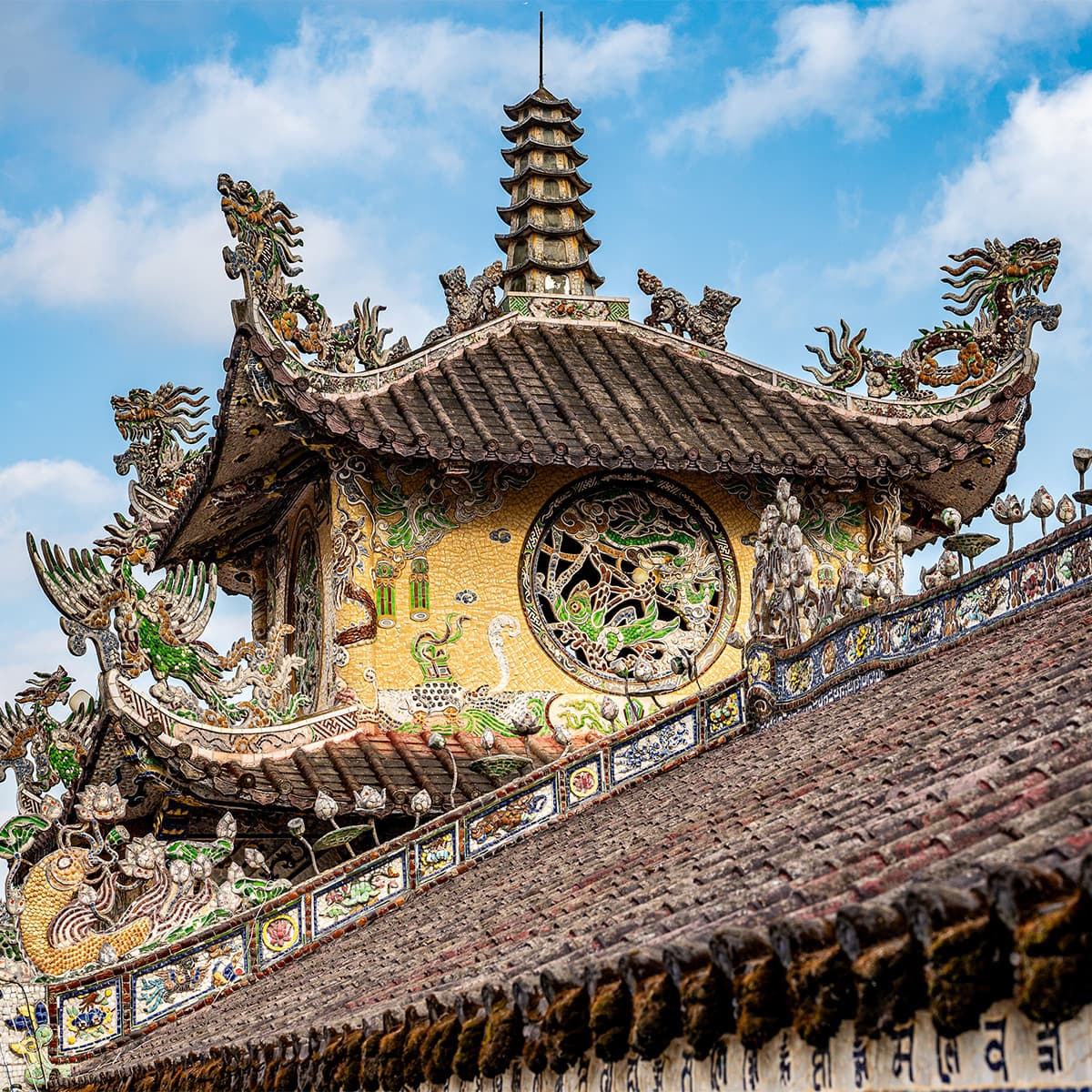 Крыша главного зала Linh Phuoc в Далате с изогнутыми карнизами и декоративными драконами, где мозаика из стекла и фарфора мерцает на солнце.